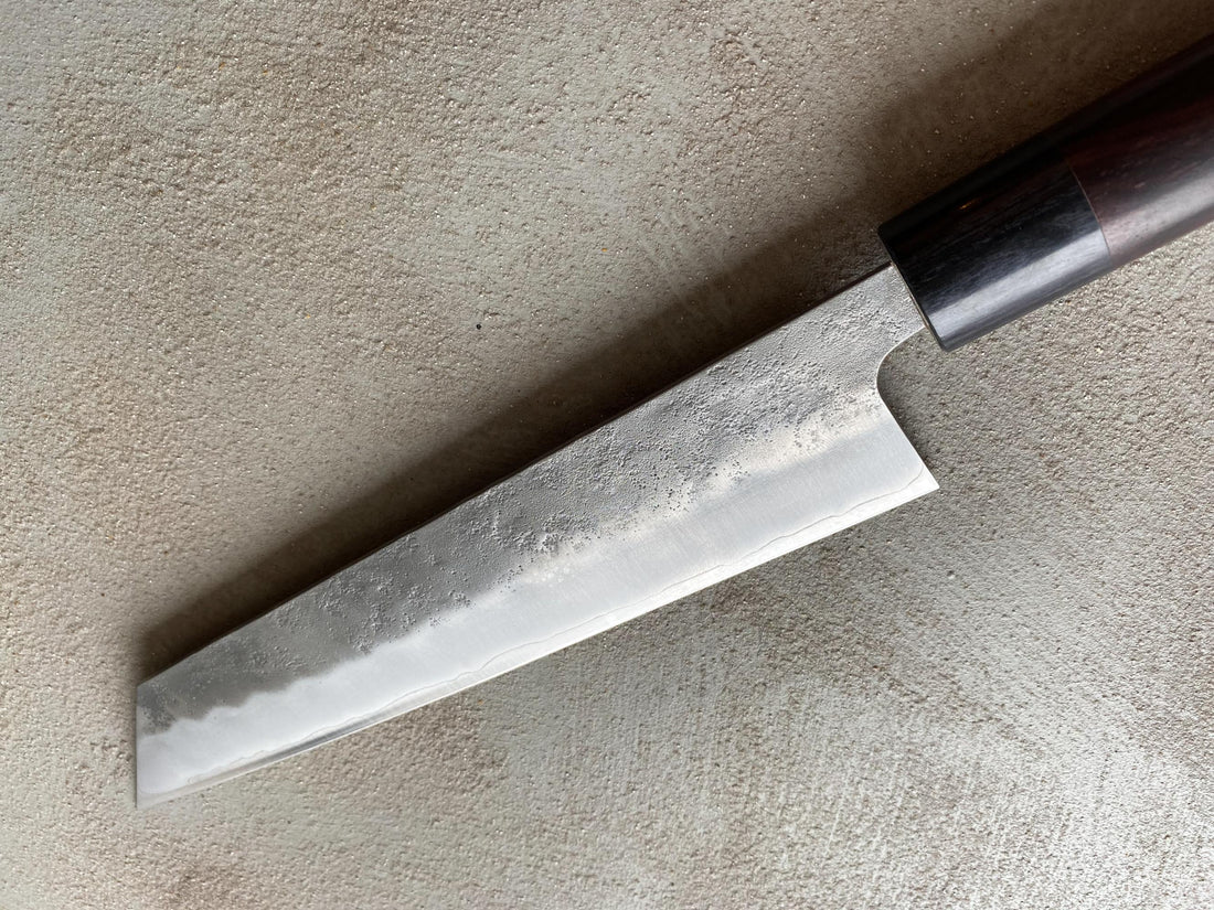 Hokiyama Kiritsuke Ginsanko Rosewood 210 mm_5  Čeprav je profil rezila Hokiyama Kiritsuke Ginsanko Rosewood nekoliko bolj raven kot pri Gyuto nožih, gre še vedno za večnamenski nož, primeren za vsakodnevno uporabo, saj je dolžina rezila 210 mm optimalna velikost za sekljanje zelenjave ter rezanje večjih kosov mesa in rib. Nož zaznamujejo odtisi kladiva nashiji.