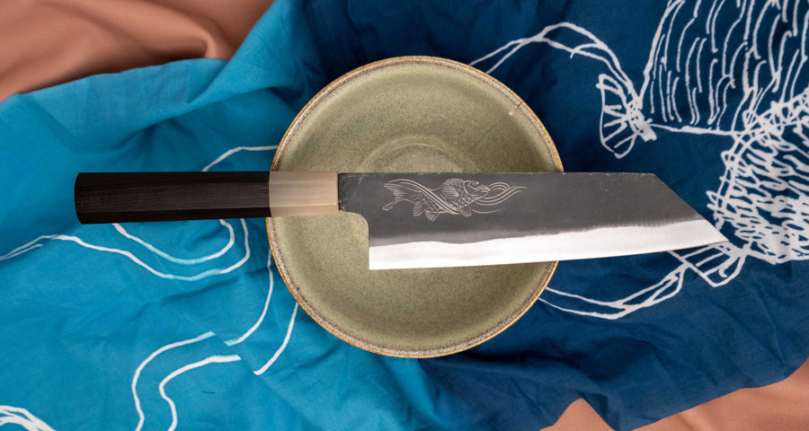 Yoshida Bunka SUJ-2 Kuro-uchi 190 mm ročno izdelan kuhinjski nož iz serije Yoshida Hamono, s trpežnim SUJ-2 jeklom, rustikalnim kuro-uchi zaključkom, ročajem iz ebenovine ter unikatno gravuro koi krapa, idealen za raznolika kuhinjska opravila in kot darilo.