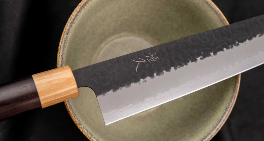 Tsunehisa Kiritsuke AS Kuro-uchi 240 mm je japonski nož, ki ga poleg enega najboljših jekel za kuhinjske nože in popolnega profila rezila (zelo tanko rezilo!), zaznamuje tudi unikatna kombinacija temne patine in odtisov kladiva, ki ju vizualno lepo dopolnjuje klasičen japonski ročaj iz palisandra in javorjevega lesa.